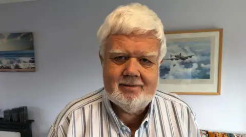 Steve Williams looking at the camera. He has white hair and a white beard. He is wearing a white shirt with blue stripes. In the background hanging on the wall is a print of a Lancaster bomber in flight.