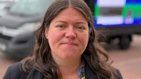 Close up of Merseyside police and crime commissioner Emily Spurrell who has long brown hair and wearing a lime green dress with red and white flowers and a grey blazer. She is standing near New Brighton beach. She is smiling.