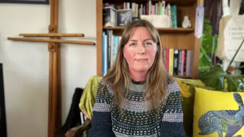 Artist Rebecca Farkas sits on a cushioned sofa in her home studio. An easel rests against the wall behind her, next to a bookcase and house plant. Rebecca has long, brown hair and wears a patterned woollen jumper. 