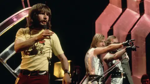 Bill Oddie singing in a yellow T-shirt in the foreground on Top of the Pops, with the other Goodies to one side in silver outfits, pointing at the crowd