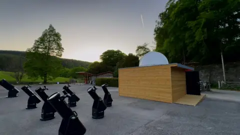 The observatory's telescopes. Seven 8in (20cm) telescopes point towards the sky. The stand in front of its robotic telescope which is contained in a wooden room with its own protective dome.