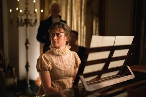 A woman looking to her right hand side, sits at a piano wearing a beige dress.