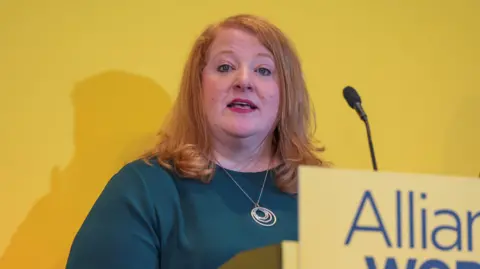 A woman with red hair is standing in front of a microphone. She is wearing a green top and a large silver pendant is round her neck. A sign saying 'Alliance' is in front of her microphone.