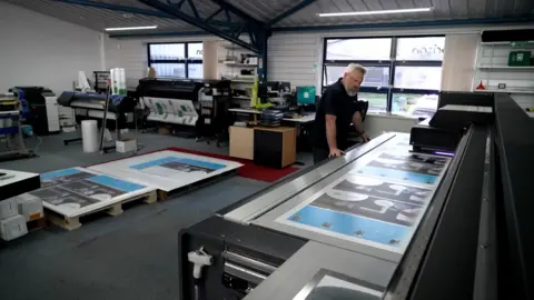 Andy Meeson/BBC A man wearing a dark blue t-shirt looks onto the printing bed of a large industrial-sized printer. He is leaning on the printer with both arms outstretched. He has a concerned look on his face, has a full greying beard and grey hair. The printer is several metres wide and appears to be part-way through printing materials. The image is taken inside a commercial facility, with dark carpets, strip lighting and white walls. Other, smaller printers can be seen in the background. There are also two wooden pallets on the ground, with printed materials on top.
