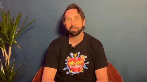 A picture of James sitting in a dark red chair in front of a dark blue wall, next to a plant on the left. He wears a black shirt with a colourful Hip-Hop Healing graphic.