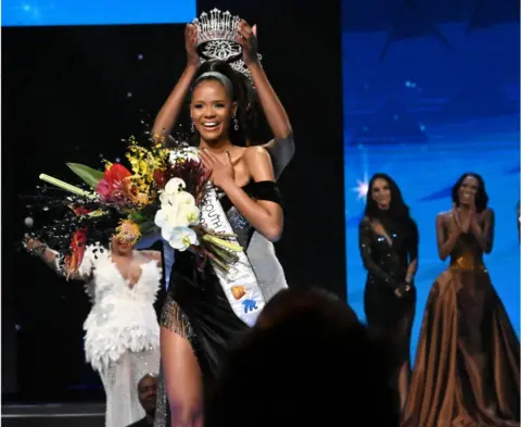 Getty Images Miss SA Ndavi Nokeri smiling in a sash with flowers as she received her crown. There are women behind her clapping.