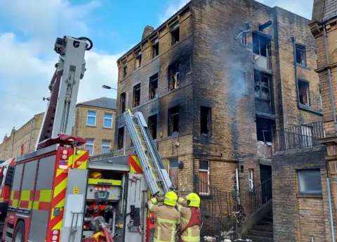 WYFRS Two firefighters pushing a ladder from a fire engine in front of a burnt out building