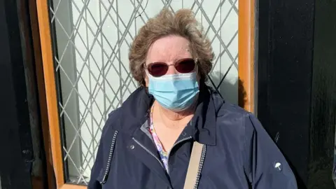 Andy Mitchell/BBC Elizabeth Lamb standing in front of a house window. She is wearing a navy blue coat and has short brown hair and sunglasses. She is wearing a blue face mask