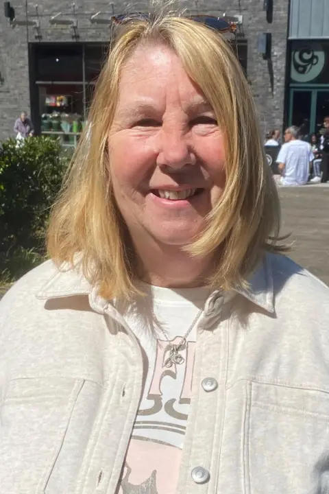 Liz Thorne, looking at the camera. She has blonde hair and is in front of a set of shops. It is a head and shoulders shot.