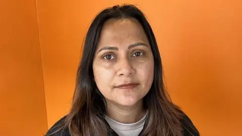 Phil Harrison/BBC A woman with long, brown hair. She is wearing a white shirt under a black jumper. She is standing in front of an orange background.