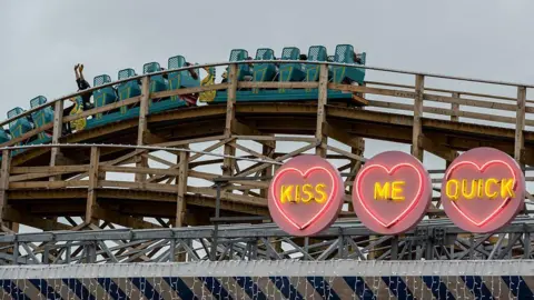 Getty Images A person on a blue rollercoaster carriage with their arms up as the cars go over a wooden arc. In front of the rollercoaster is three neon heart lights. With in each heart are the words "kiss", "me" and "quick".