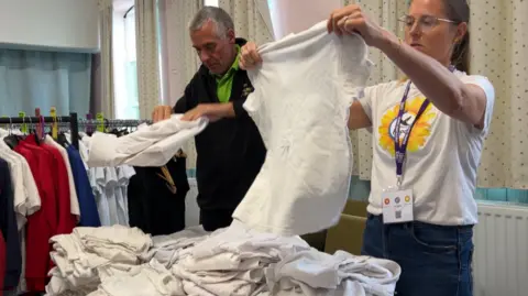 Volunteers sorting through the uniforms. A man with short grey hair and wearing a green t-shirt underneath a black jumper is folding a white shirt. Next to him is a woman with light brown hair in a pony tail and with glasses. She is holding up a white shirt. In front of them is a pile of white shirts and to the side is a clothes rail with various items on.