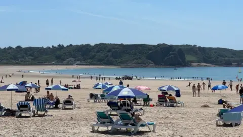 A busy beach on a summer day shows several blue and white sun-loungers and beach umbrellas. The tide is out, with many people in the shallow waves. There are also several sunbathers on the sand. In the background, green hills and trees rise above the bay.