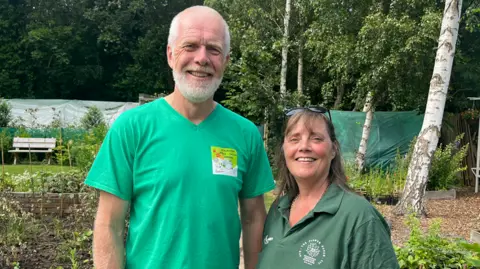 Nicky Morgan is pictured with her husband Dale. He has white hair at the sides and a white beard. He has a green t-shirt. She has long hair, a green polo shirt and black sunglasses on her head. They are standing in a park.