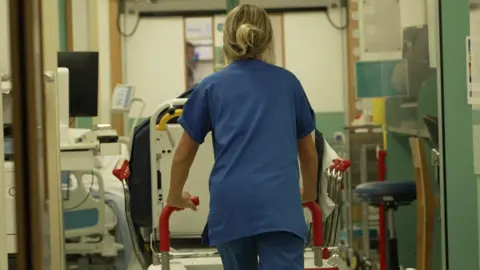 An NHS worker pushing a bed through a hallway in a hospital. She is wearing blue scrubs and has her blonde hair up in a bun. 