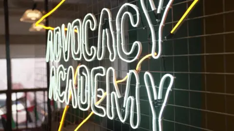 A white and yellow neon sign attached to a black tiled wall. The sign says Advocacy Academy. 
