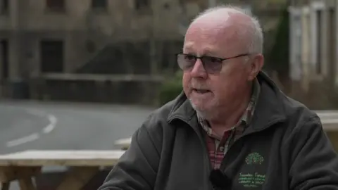 Steven Willock wears glasses and a green fleece with the Trawden Forest Community Centre logo branding on, as he sits outside on a bench.