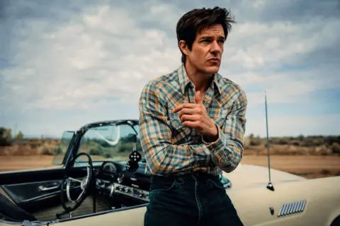 Chris Phelps Brandon Flowers in a checked shirt, leaning against a classic American convertible, with fluffy dice hanging from the rear view mirror