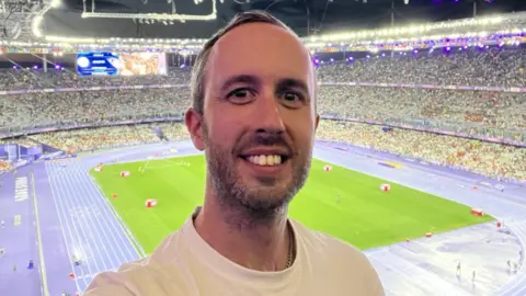 Chris Brown A photo of a man smiling in front of a stadium. The stadium is purple and the man has short brown hair and has a beard. He is wearing a while T-shirt. 