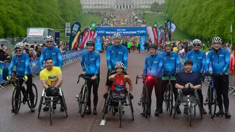 Pacemaker The picture shows three people on wheelchairs in front of other people dressed in blue on bicycles. Stormont and hundreds of marathon runners are behind them 