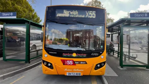 Martin Giles/BBC The front of a yellow Sanders bus. In the sign at the top it says 'Norwich express via Coltishall X55'. Either side of the bus are bus shelters.
