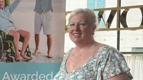 Leanne Shakespeare A woman in a pastel floral top. She is standing next to a window and a banner that has people on a beach. One person on the banner is standing and the other is in a wheelchair. The woman has short blonde hair and is smiling.