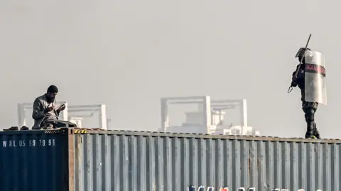 AFP A supporter of the Pakistan Tehreek-e-Insaf (PTI) party offers prayers on top of a shipping container as a paramilitary soldier chases him during a protest demanding the release of former prime minister Imran Khan, at the Red Zone area in Islamabad on November 26, 2024. 