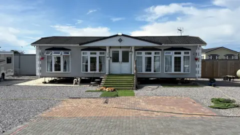 Ben Schofield/BBC A wide shot of a static caravan. It is set back from where the camera is stationed, with an expanse of block paving and a stone driveway in the foreground. A small stairway leads up to the caravan's double, uPVC-type doors. Either side of the doors are two sets of windows, meaning across the front there are four sets of windows in total. The caravan is symmetrical and imposing against a light blue, partly cloudy sky. 