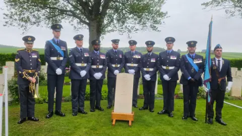 Crown Copyright 10 men in RAF uniforms, one with a bugle, stand in front of a grave stone