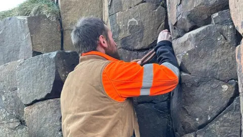 National Trust A man in work clothing uses told to remove coins from a rock. he is standing with his back to the camera