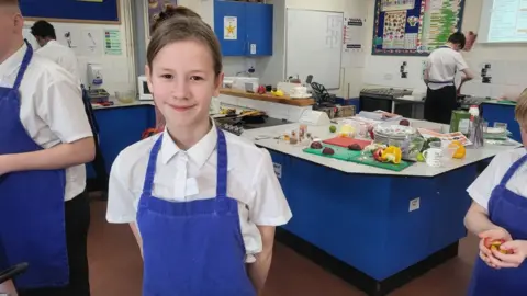 Amelia stands with her hands behind her back in a kitchen classroom. She is smiling slightly at the camera and wearing a white shirt and bright blue apron. 