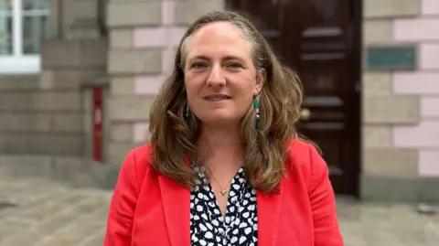 Hilary Jeune is smiling at the camera wearing a blouse underneath a red suit jacket. She is standing outside the government building which is blurred.