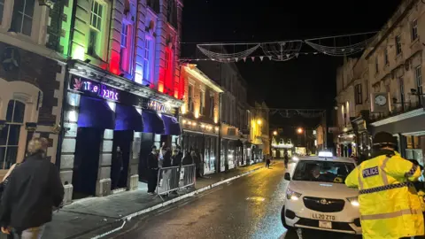 A night scene on a busy street with a police officer in the corner and people waiting outside a venue