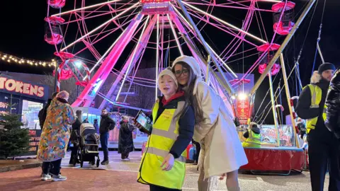 BBC/Kian Boyle A woman wearing white winter clothes is hugging a small boy wearing a winter hat form behind