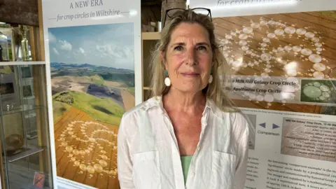 Monique is standing at her exhibition in front of some posters showing crop circles. She is wearing a white blouse and is looking at the camera.