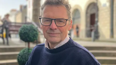 Barrie Duerden is facing the camera in a navy blue jumper with shirt collar visible under. He is wearing black-rimmed glasses and has short fair hair. Behind him, limestone coloured steps and buildings are visible but are slightly blurred. Some benches are also in the background behind his right shoulder.