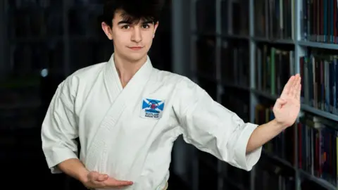 A teenager with dark hair, standing indoors and wearing a white karate gi with a small Scotland flag on it. He has one palm open, as if about to compete.