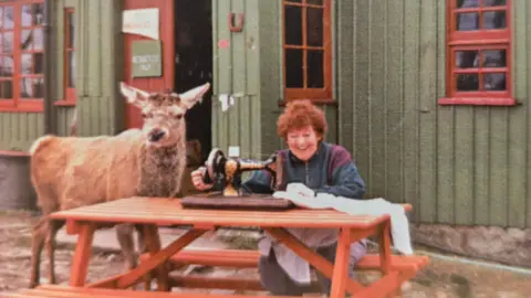 Joan Martindale/Hostelling Scotland Joan Martindale is sitting at a table outside the youth hostel working at a sewing machine. She is laughing because a deer is standing at the table next to her.