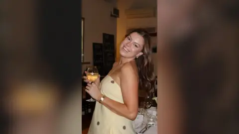 A young woman with long brown hair smiles while holding a glass of wine. She is wearing a strapless yellow dress.