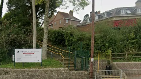Woodroffe School buildings are partially visible above a hedge as seen from the road. A white school sign is at street level.