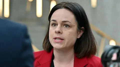 Kate forbes mid speech. She has a brown shoulder length bob and is wearing a red blazer
