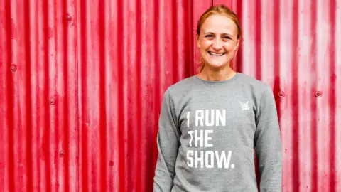 A woman wears a grey sweater against a red background that reads 'I RUN THE SHOW' in capital letters. She wears her hair up and gives a big smile. 