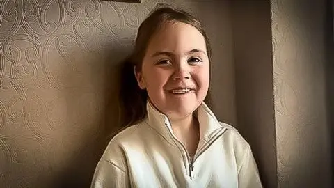 Family photo Darcy is smiling at the camera as she stands in front of a brown patterned wallpaper wall. she has long light brown hair tied back and a beige polar fleece top