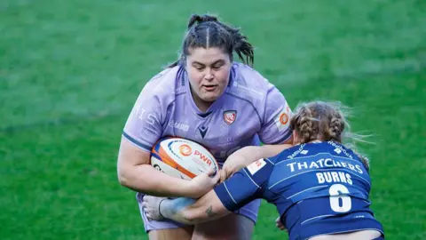 Maud Muir runs into a tackle while holding a rugby ball under her right arm to her body, as Delaney Burns (Bristol Bears) crouches and holds an arm around Muir's waist