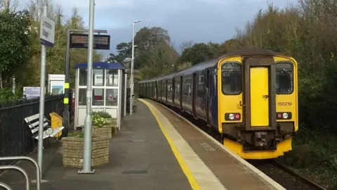 A GWR train pulls into Lympstone Village station. The train has a yellow front. The line is single tracked.