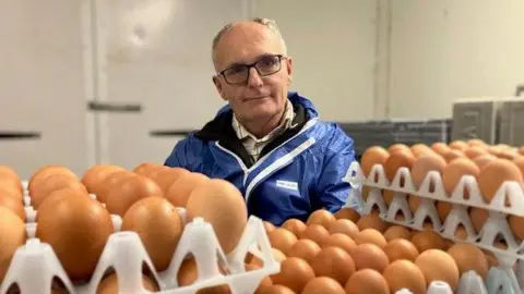 BBC/Andrew Sinclair Farmer Simon Dann sits in his egg room with boxes of eggs in the foreground 