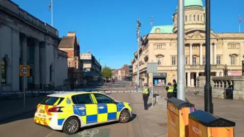 BBC A police car is parked across Carr Lane and three police officers stand in front of scene guard tape across the pavement.