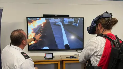 Two men, one in uniform and another in a striped shirt and wearing headset, with both men looking at screen displaying a fire scene.