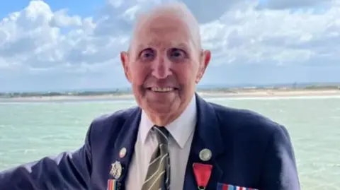 Old man in blazer with medals smiles for camera with beach and sea behind him