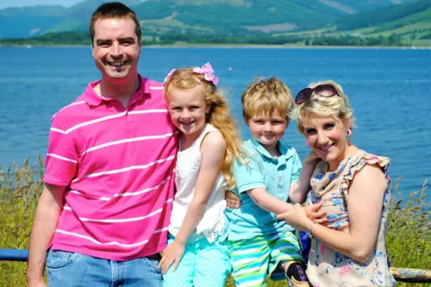 Sam Hey Susan, Lily, Hamish and Susan pictured on holiday. A large area of water behind them is blue and there are green, tree-covered hills on the opposite shore.